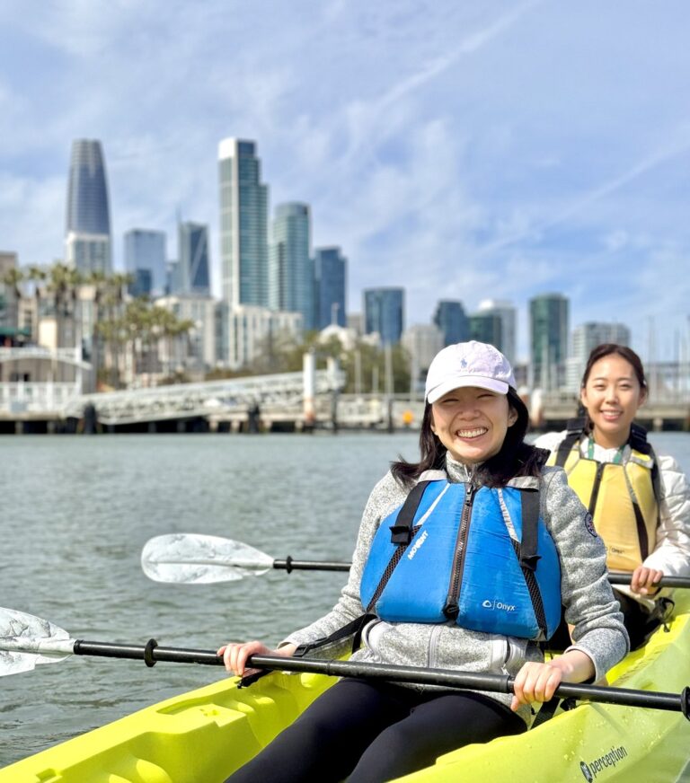 Two young paddlers in a kayak with the downtown skyline behind them.