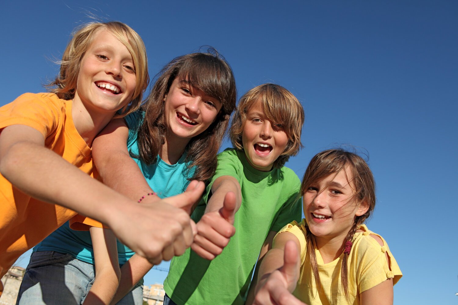 Four children in bright t-shirts grinning and giving a thumbs-up against a clear blue sky.