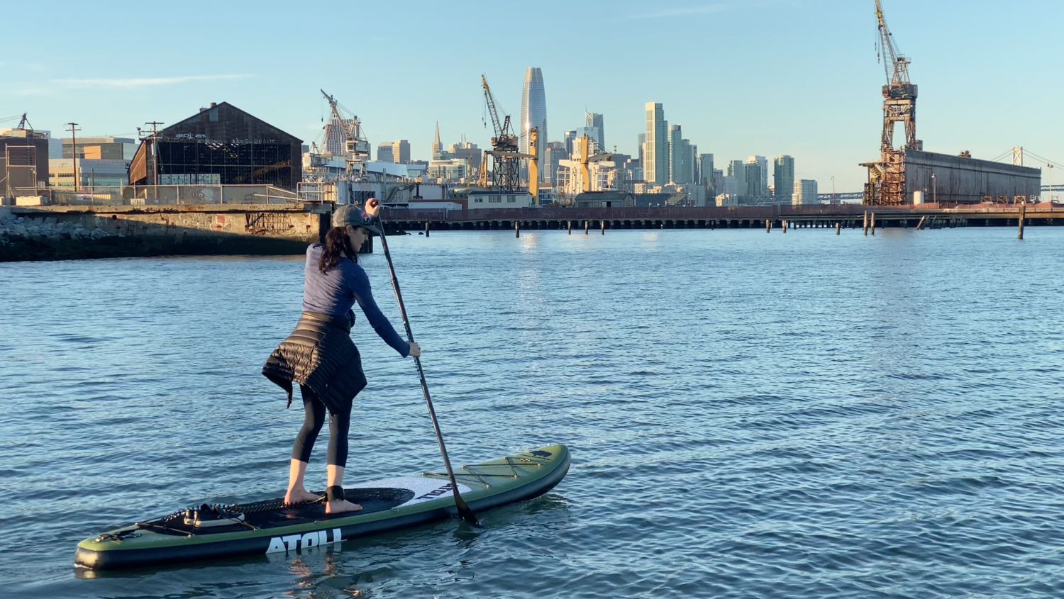 A paddler crossing flat water on a SUP with the downtown skyline and shipyard cranes behind.
