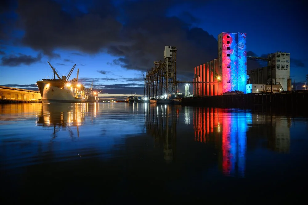 The waterfront at night, the cranes lit and reflected in still water.