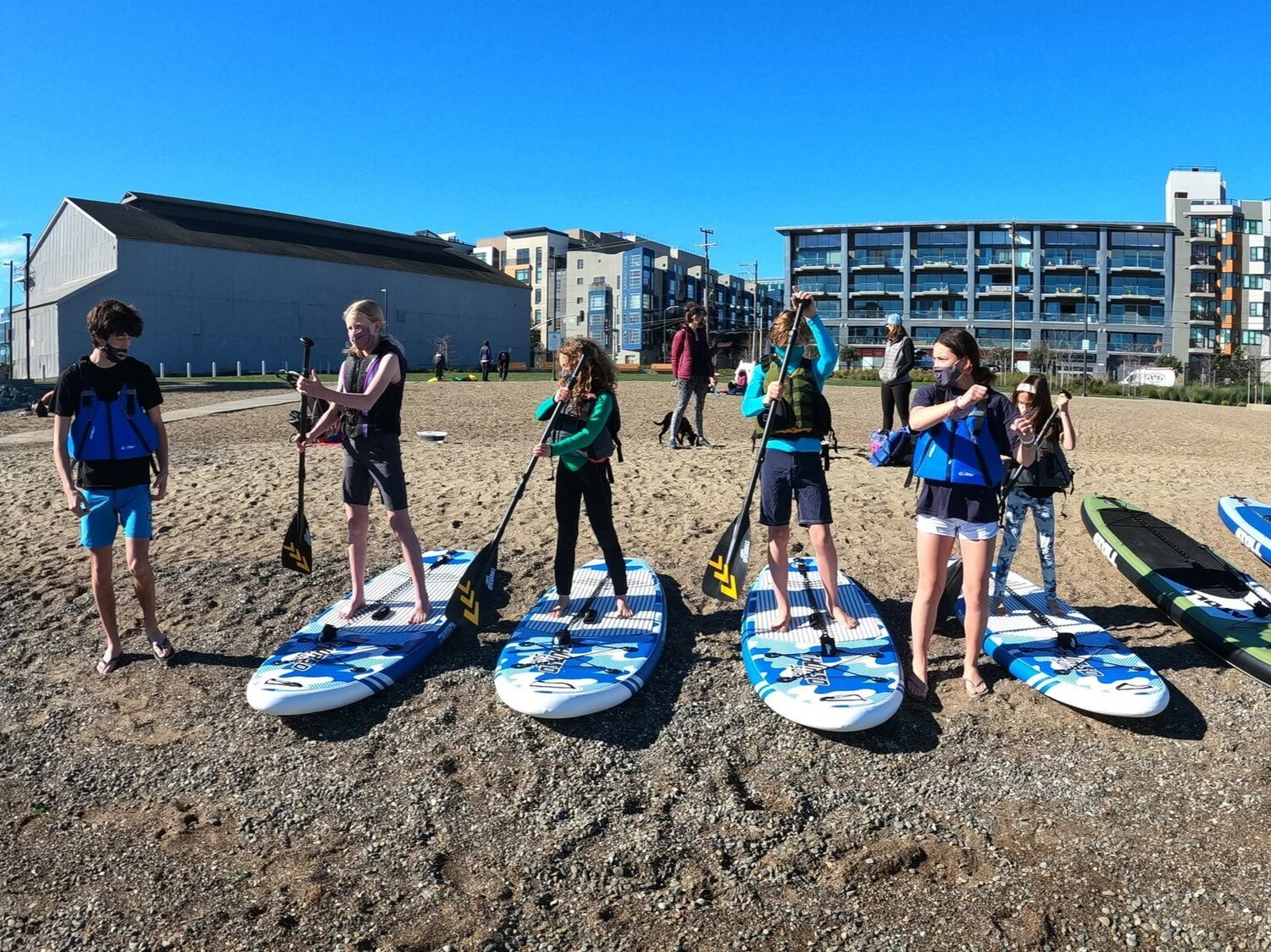 A group of young paddlers standing on their boards on the gravel beach, ready to launch.