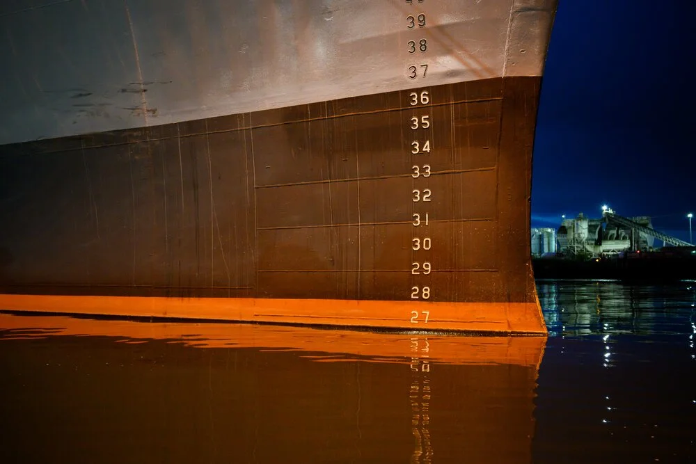 The side of a ship's hull at dusk, draft numbers painted up the steel above an orange boot stripe.