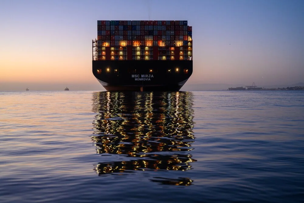 A lit container ship lying at anchor at dusk, its lights reflected on flat water.