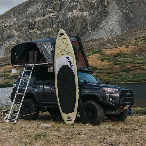 Two paddleboards leaning against a four-wheel drive with a roof tent, below a rock face.