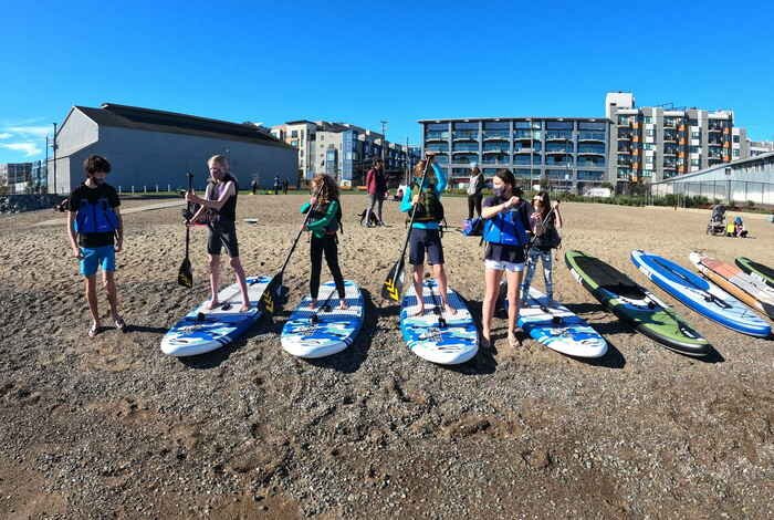 Young paddlers standing on boards laid out along the gravel beach, apartment blocks behind them.