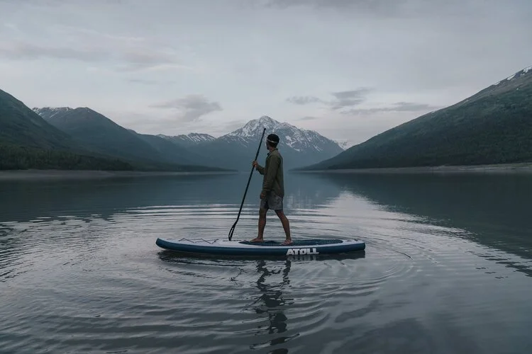 A paddler crossing flat water below steep mountains under low cloud.