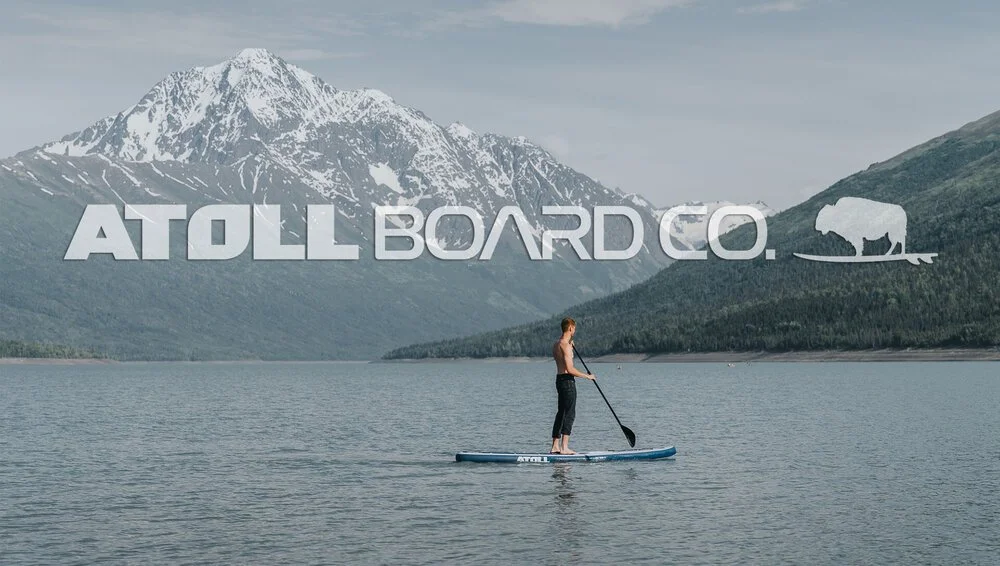 A paddler on a mountain lake beneath a snow-covered peak, with the Atoll Board Co wordmark set across the sky.