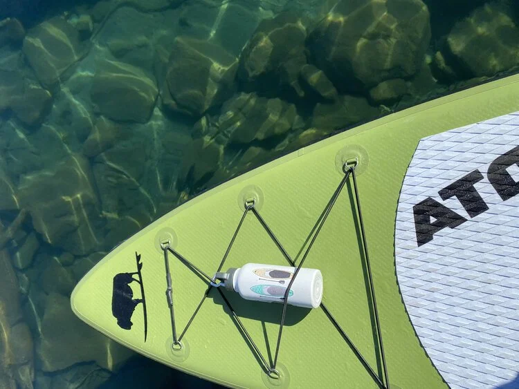 A green inflatable board floating on clear water, a bottle stowed under its bungee.