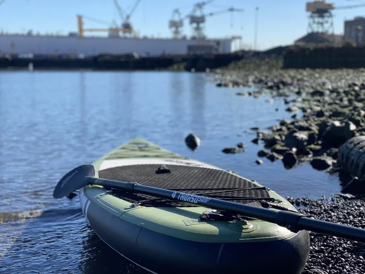 An inflatable board resting on the pebble beach with the shipyard cranes behind.