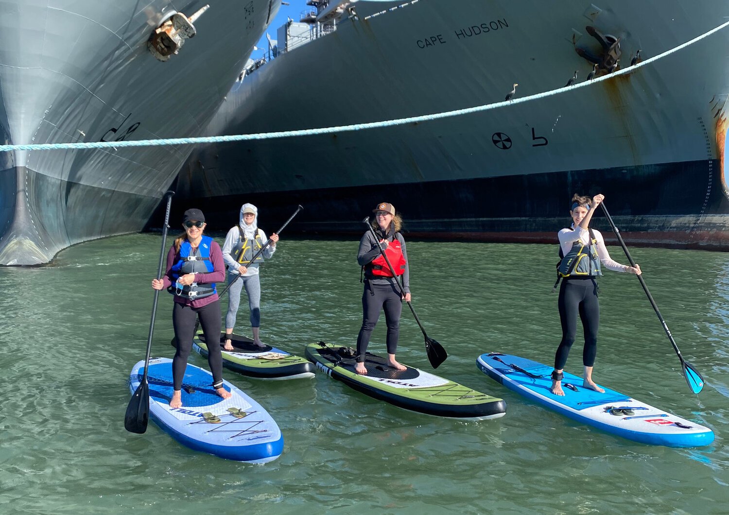 Four paddleboarders in buoyancy aids lined up on their boards beneath the bow of a moored ship.