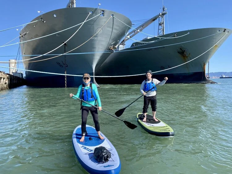 Two paddleboarders passing in front of the bows of two moored ships.