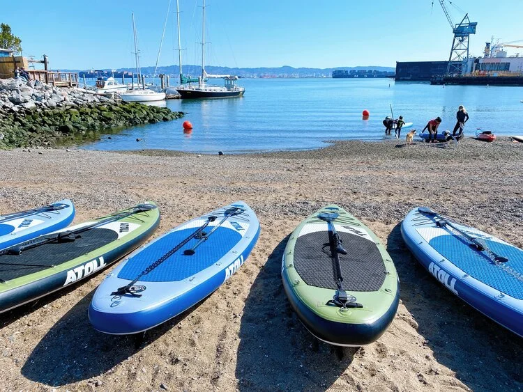 Inflatable boards lined up on the beach with moored yachts and a crane behind.