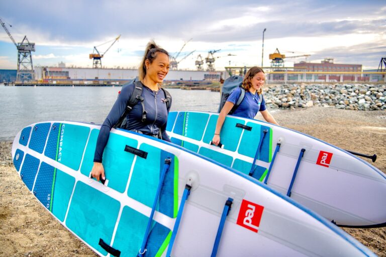 Two people carrying paddleboards up the beach, the cranes behind them.