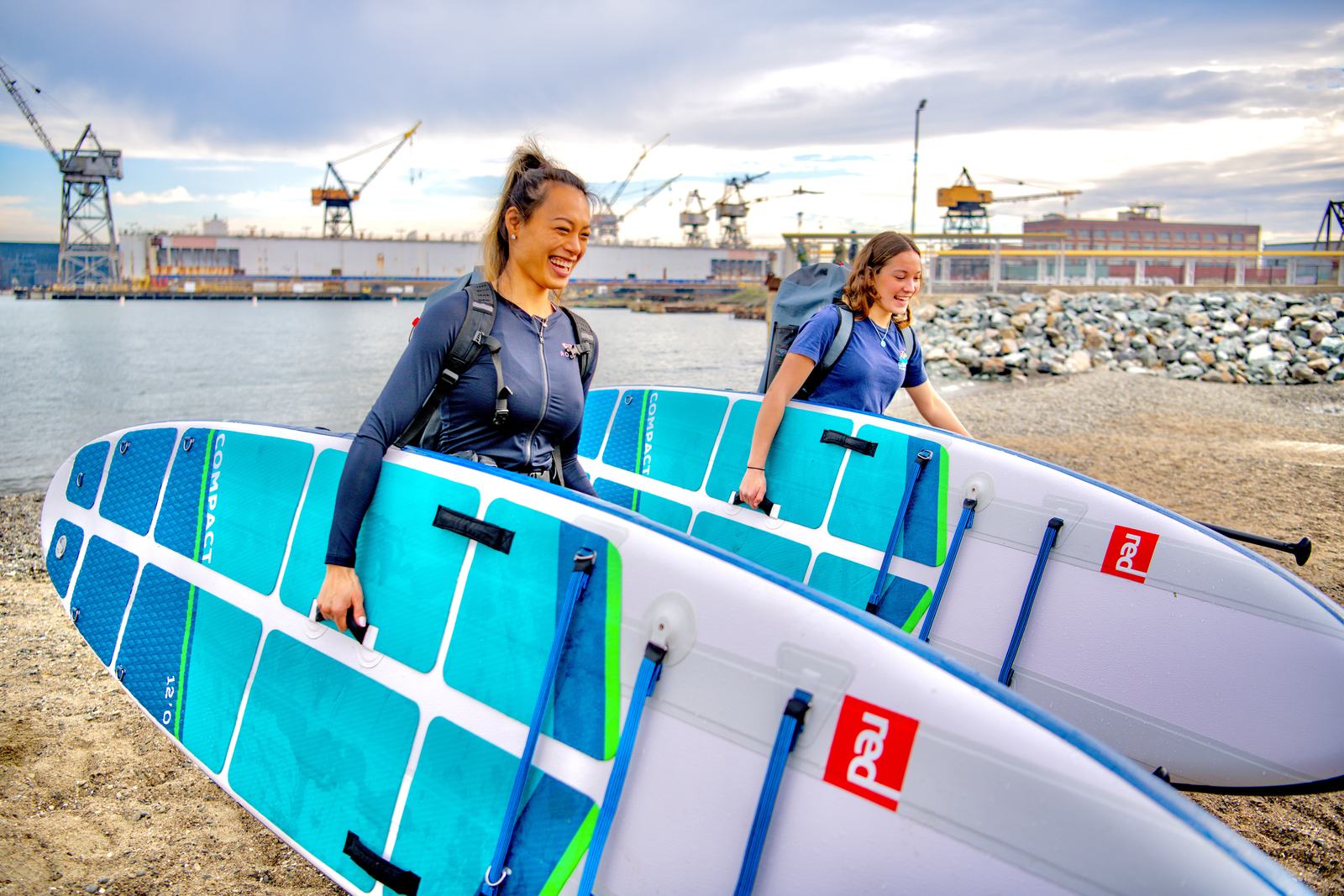 Two people carrying paddleboards up the beach, the cranes behind them.