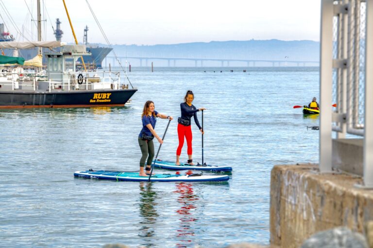 Two people on inflatable paddleboards on flat water beside the pier.