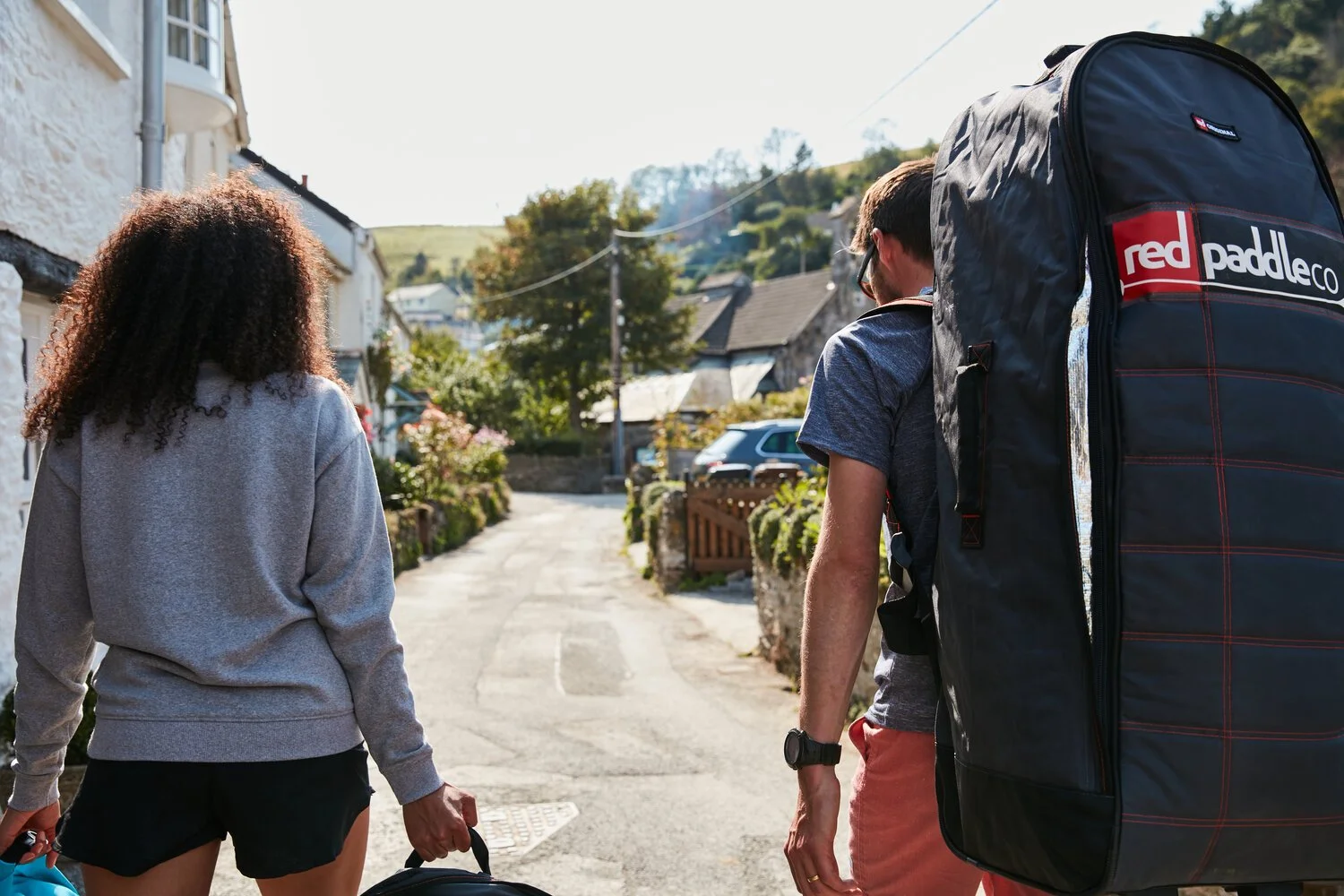 Two people walking down a village lane carrying a paddleboard in a wheeled backpack.