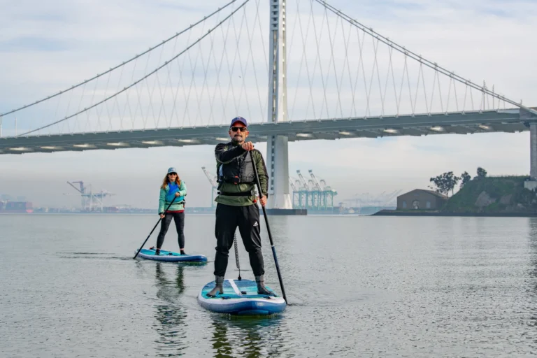 Two paddlers on boards in front of the Bay Bridge in flat winter light.