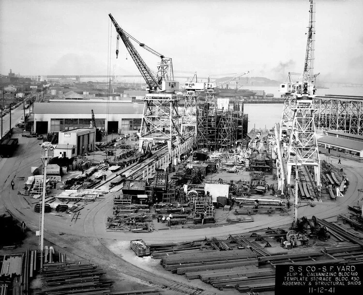 A black-and-white photograph of the shipyard mid-construction, cranes standing over a part-built hull.