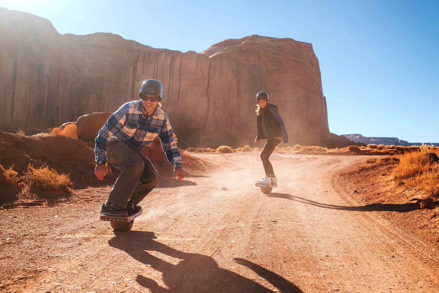 Two riders on Onewheel boards on a dirt track between red sandstone buttes, long shadows in low sun.