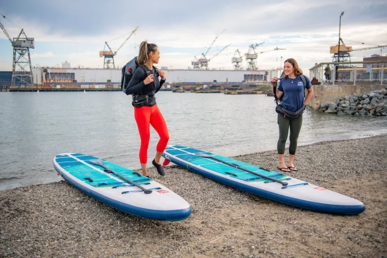 Two paddlers standing on the beach beside two inflatable paddleboards, the Pier 70 shipyard cranes behind them.