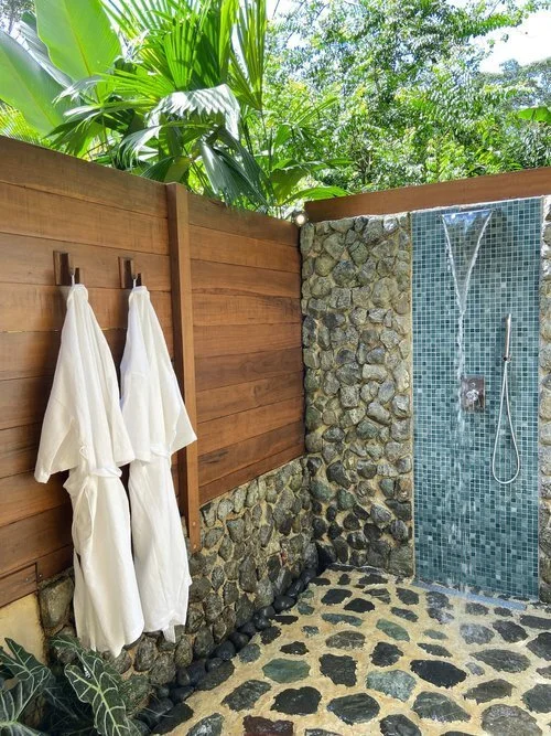 An open-air shower with white towels hanging on a timber wall and a pebbled floor.