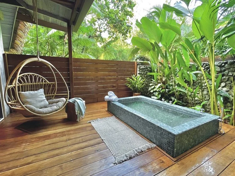 A hanging rattan chair and a stone plunge bath on a timber deck backed by banana leaves.
