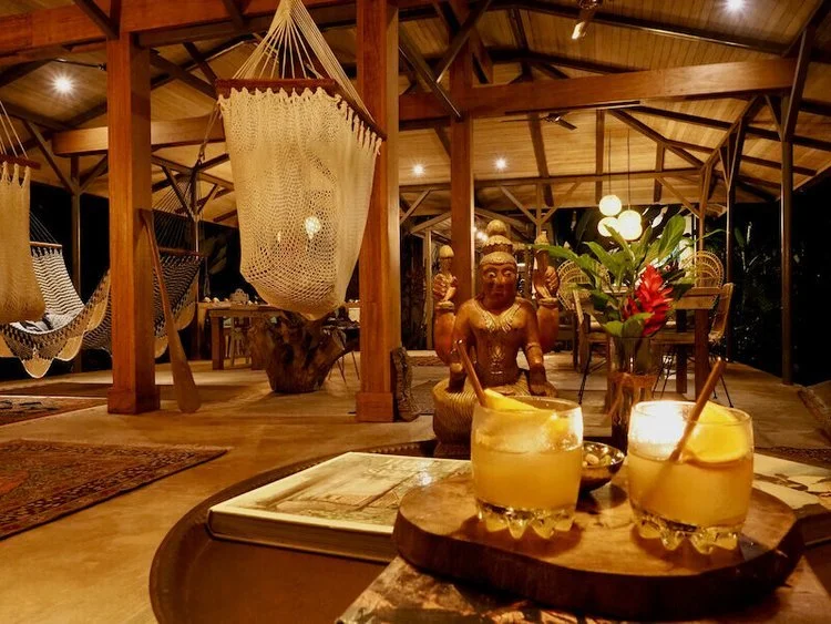 Two orange cocktails on a low table in a lamplit open-air lounge hung with hammocks.