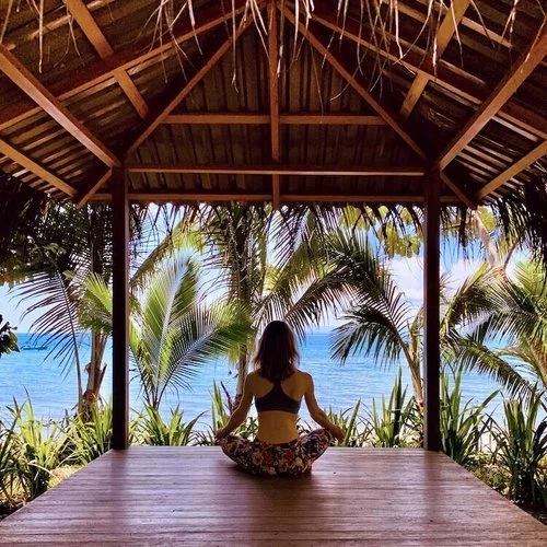 A woman sitting cross-legged in an open-sided timber pavilion, looking out through palms to the sea.