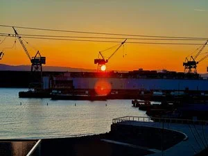 The sun setting behind the shipyard cranes, low over flat water.