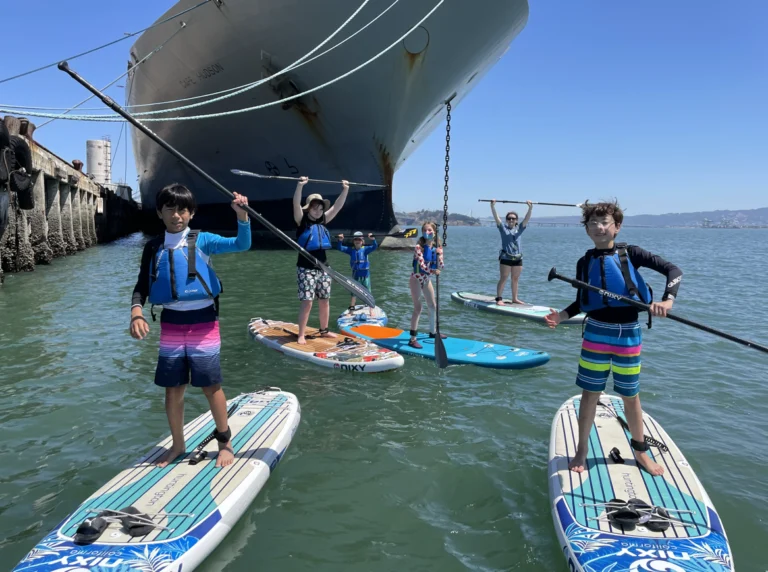 Campers on paddleboards raising their paddles under a ship's bow.
