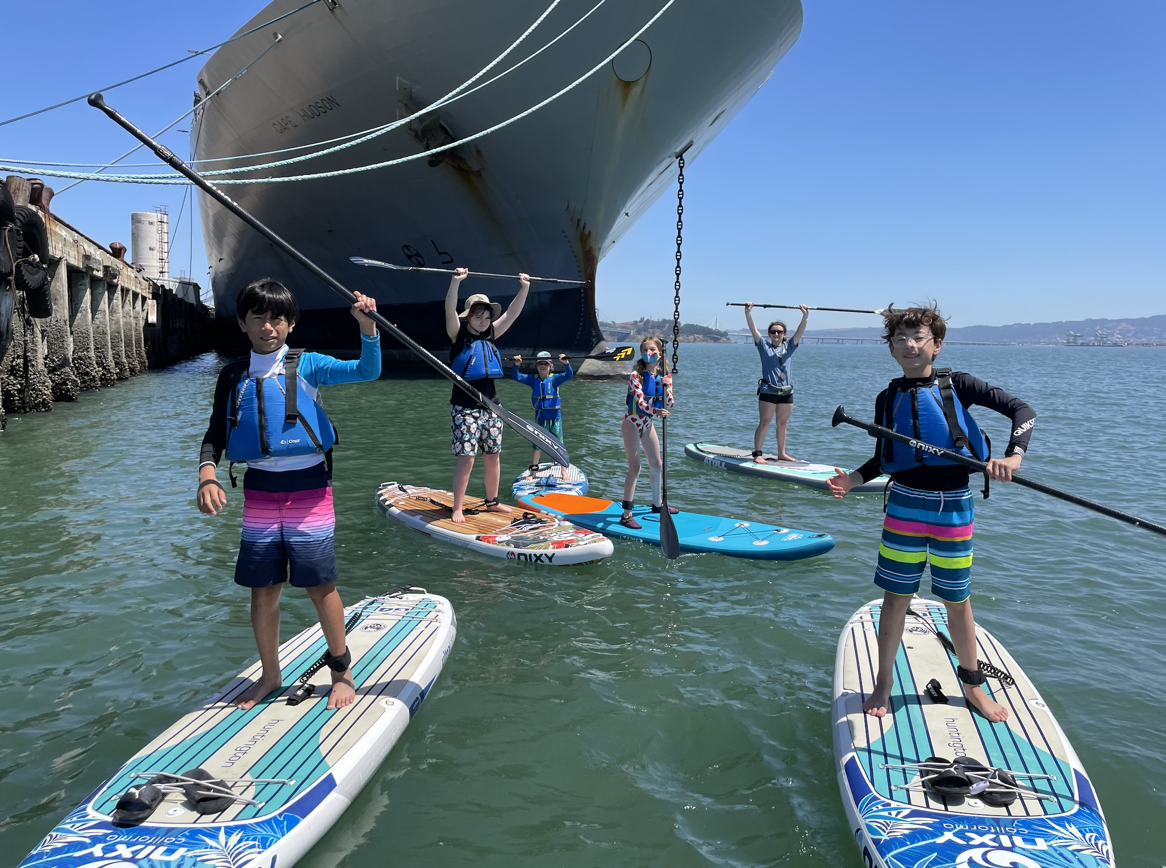 Campers on paddleboards raising their paddles under a ship's bow.