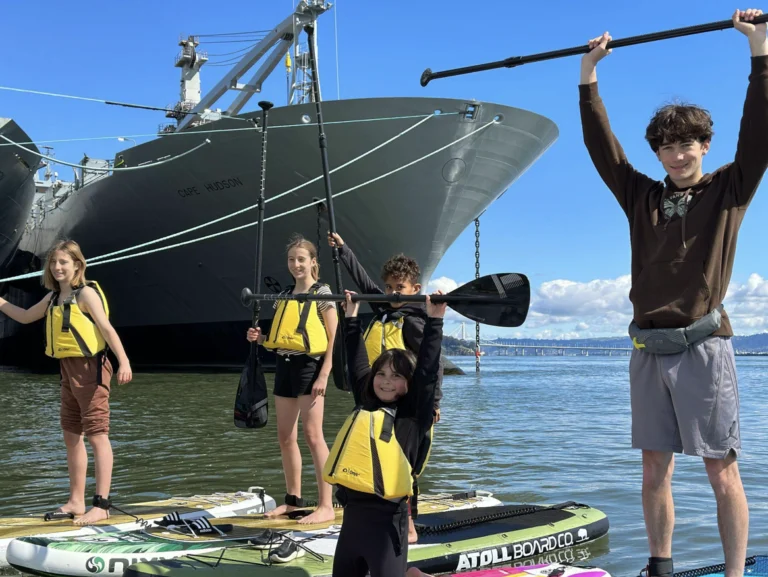 Campers in life jackets on the water beneath the bow of a large grey ship.