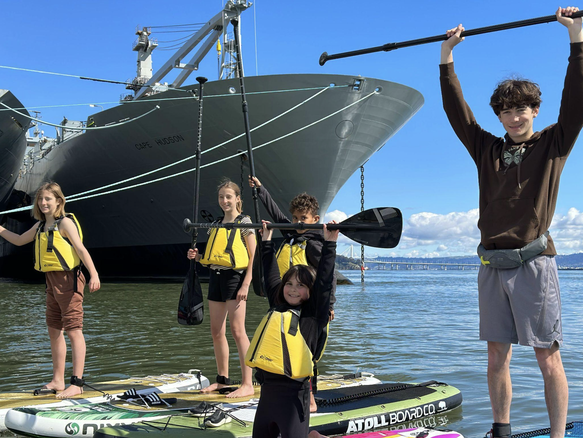 Campers in life jackets on the water beneath the bow of a large grey ship.