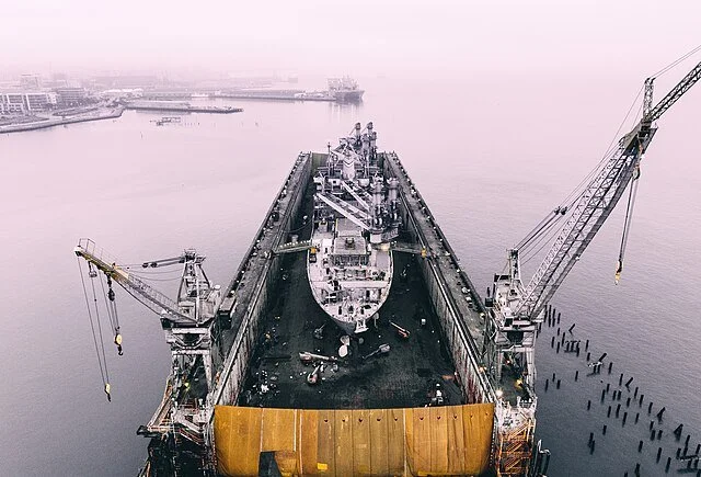 A ship sitting high in a floating dry dock with its hull exposed, cranes standing alongside.