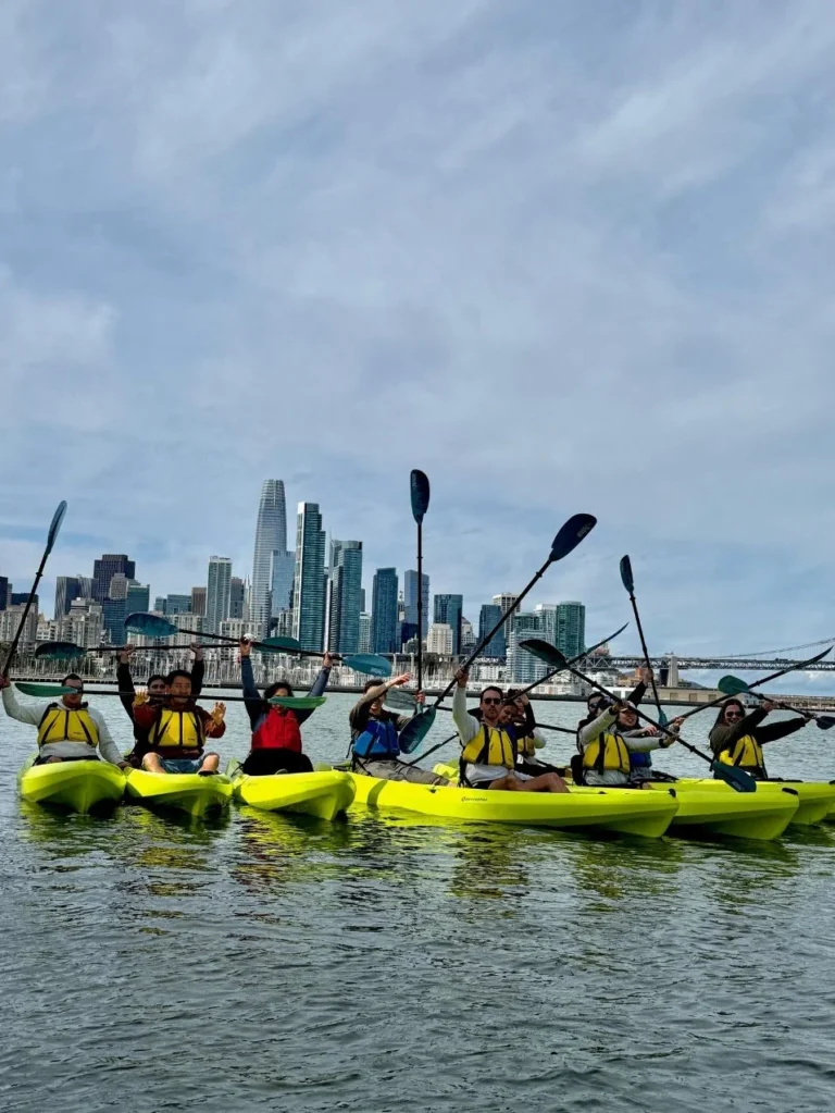 Kayakers rafted together on the water with the downtown skyline behind.