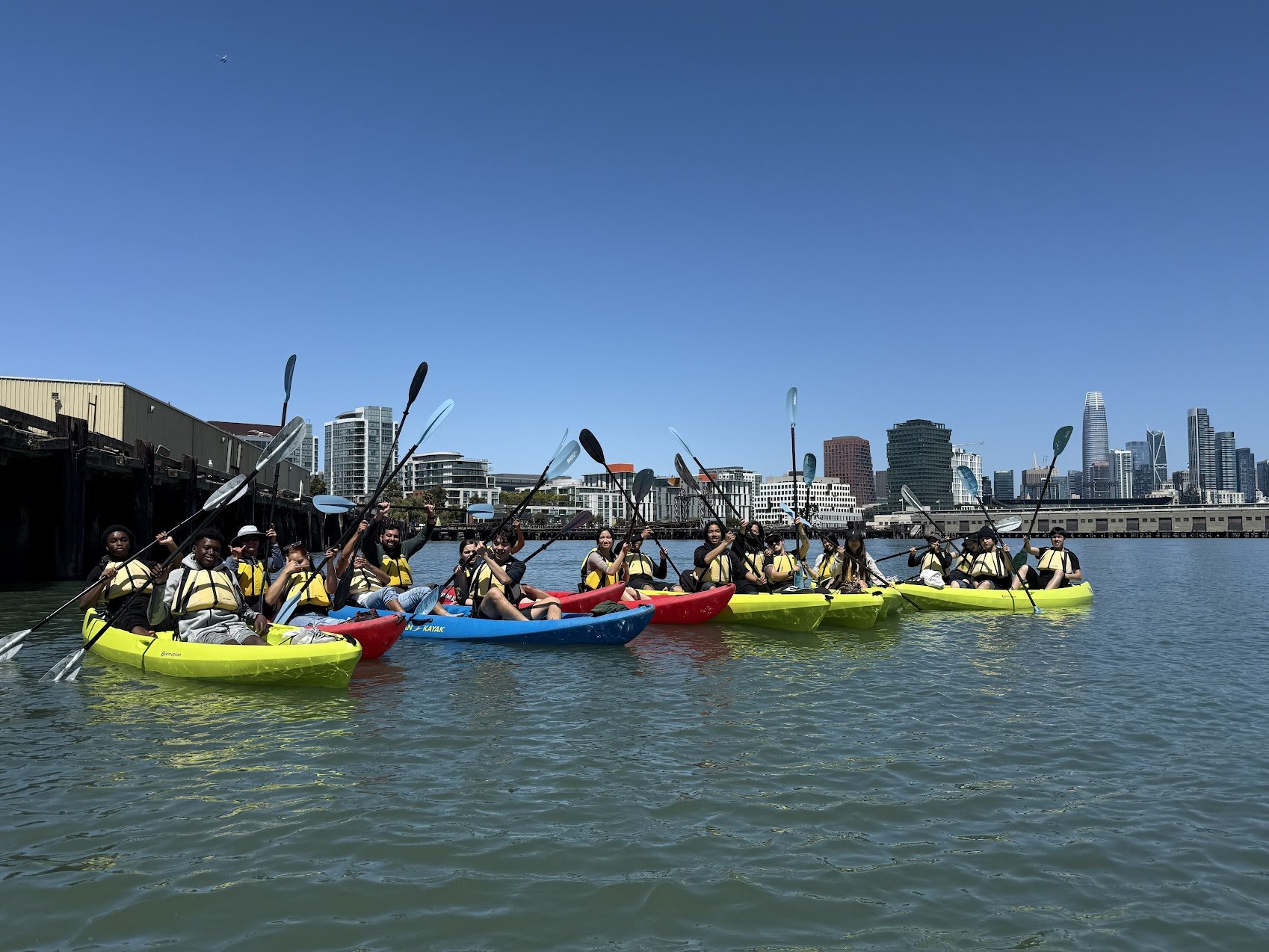 A line of yellow tandem kayaks setting off along the waterfront with the downtown skyline behind.