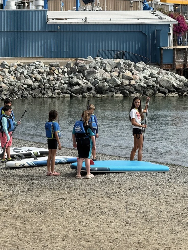 Children in life jackets on the beach beside a paddleboard.