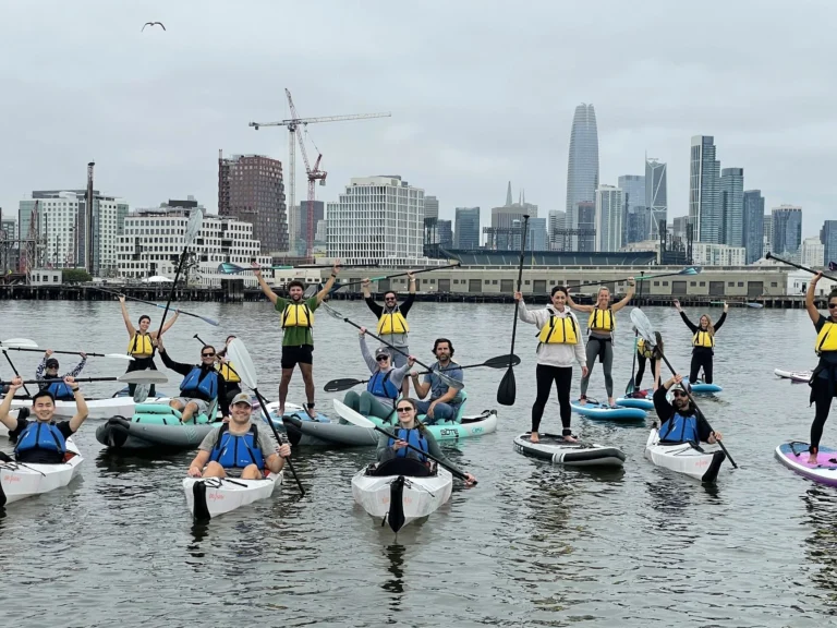A large group of paddlers on the water, cranes and the skyline behind.