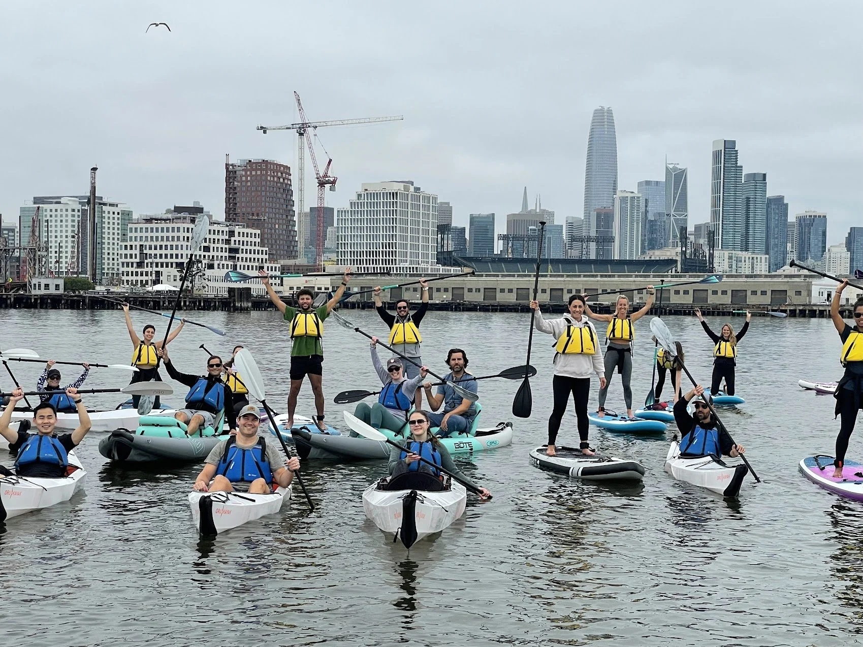 A large group of paddlers on the water, cranes and the skyline behind.