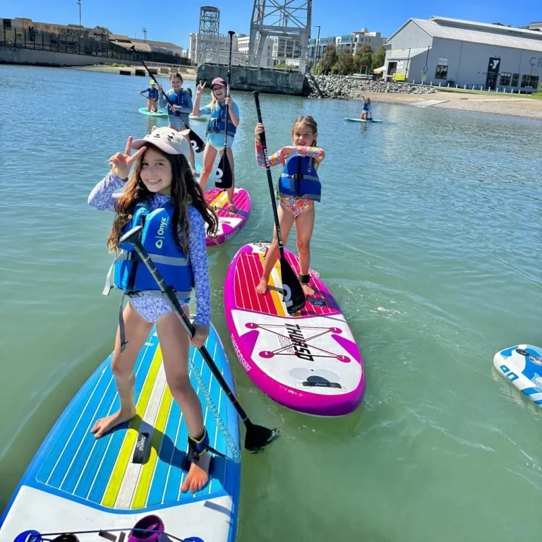 Young campers on bright paddleboards on the water at Crane Cove Park.