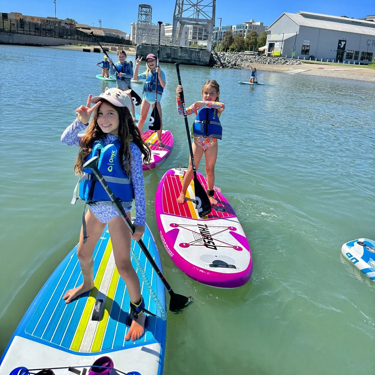 Young campers on bright paddleboards on the water at Crane Cove Park.