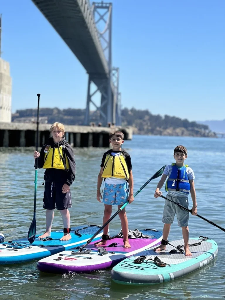 Three young paddlers standing on paddleboards under the Bay Bridge.
