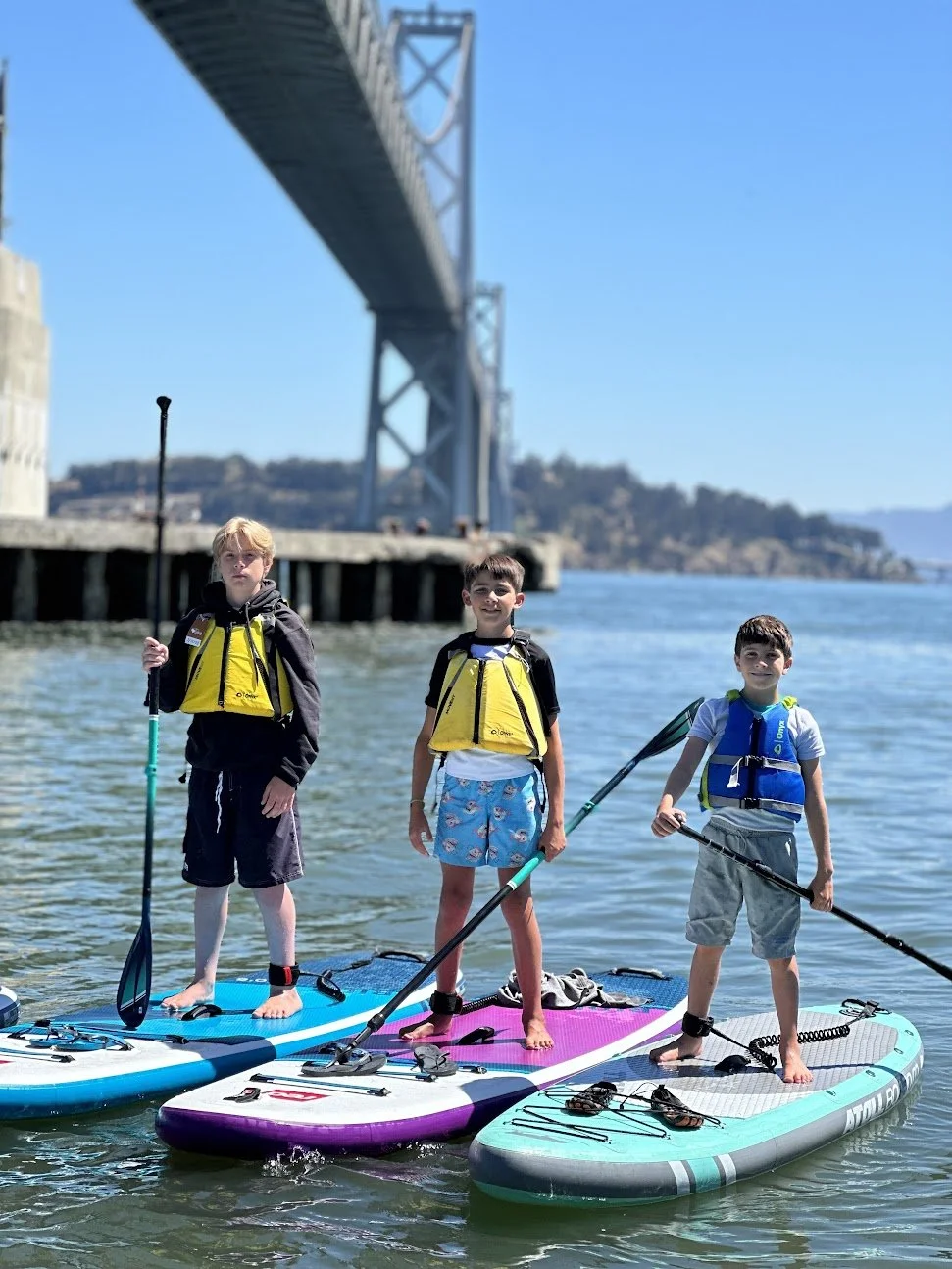 Three young paddlers standing on paddleboards under the Bay Bridge.