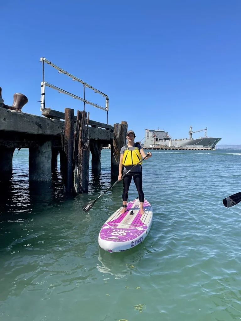 A paddler on a pink board beside a pier, downtown San Francisco behind.