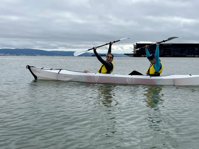 Two people in a white sea kayak holding their paddles above their heads.