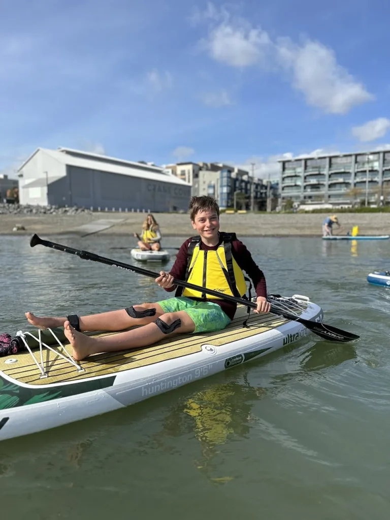 A young paddler sitting on a paddleboard with legs stretched out, the Crane Cove shed behind.