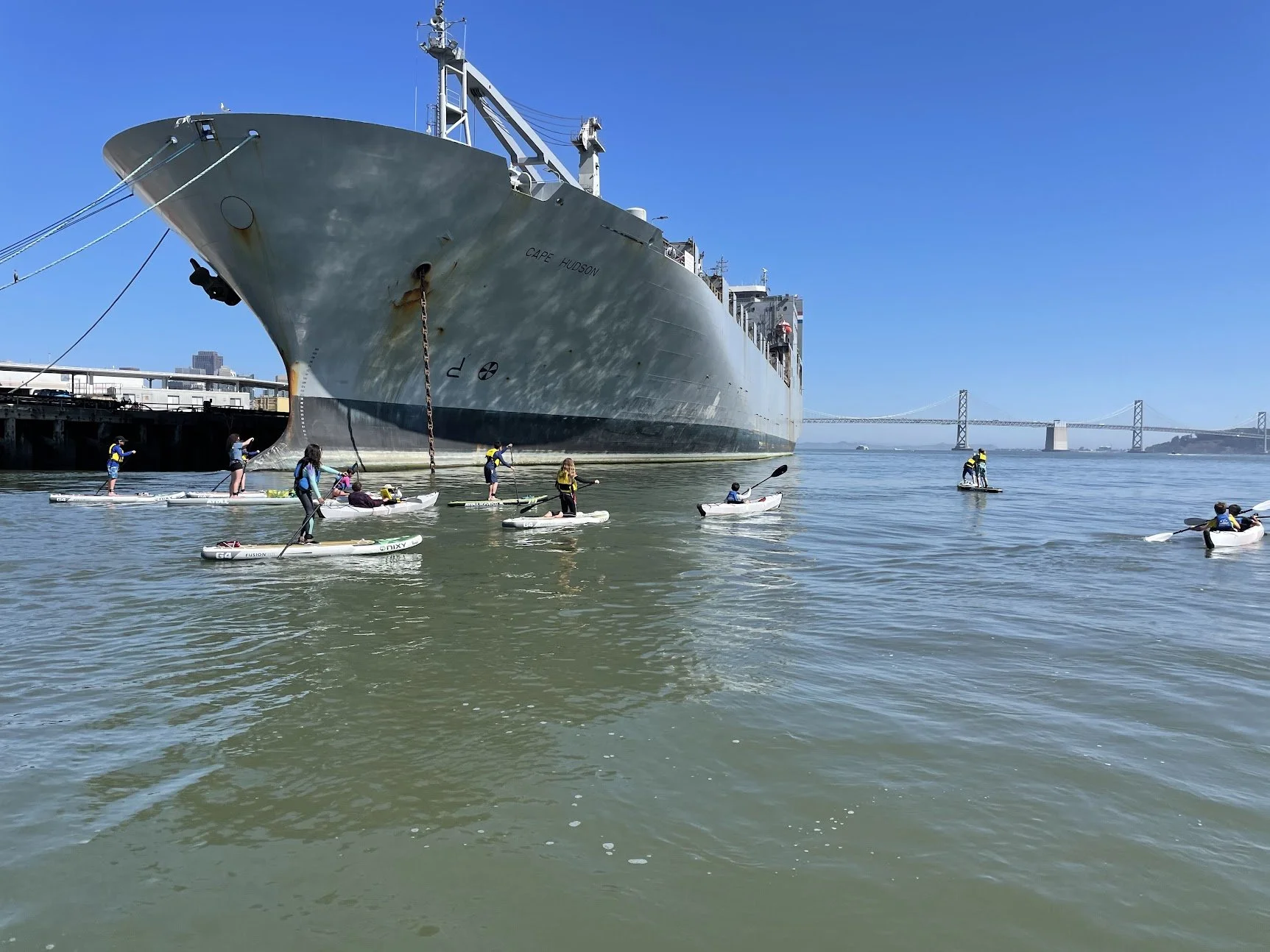 Kayakers passing beneath the bow of a large grey ship with the Bay Bridge beyond.