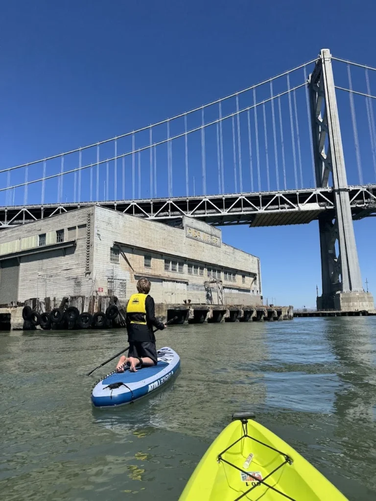 A paddleboarder under the Bay Bridge, seen over the bow of a yellow kayak.