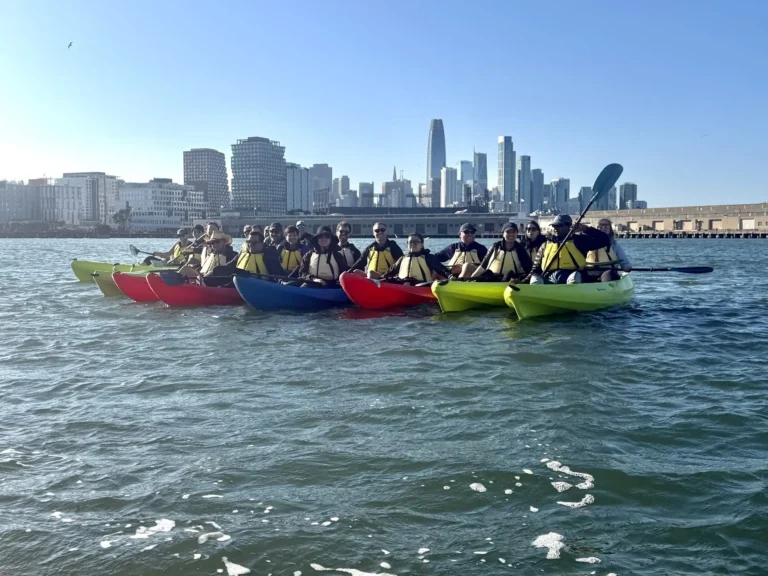 A line of kayaks on open water with the downtown San Francisco skyline behind.