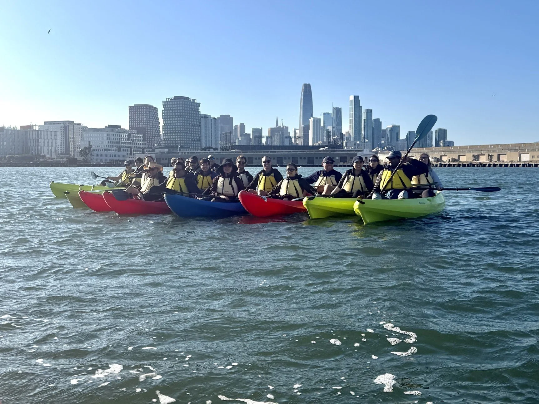A line of kayaks on open water with the downtown San Francisco skyline behind.